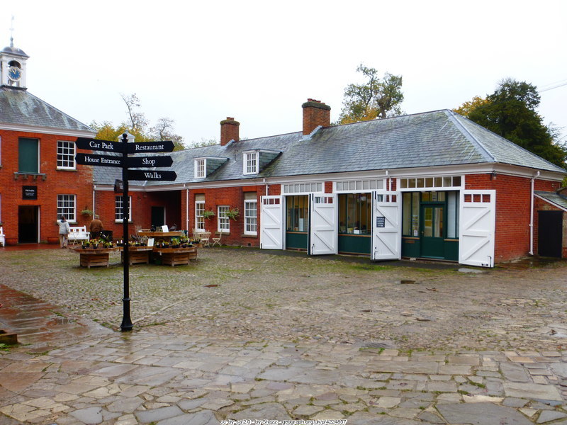Old stable yard with a black signpost at Hatchlands Park, in a stone flagged courtyard of traditional red brick buildings with grey slate roofs, white painted stable doors open to reveal rooms converted inside (cc-by-sa/2.0 - © Shazz - geograph.org.uk/p/4224857)